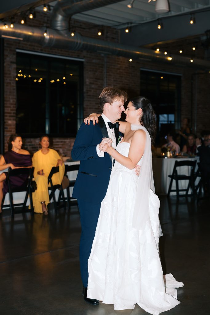 Caroline and Josh share their first dance in industrial venue under string lights with guests watching
