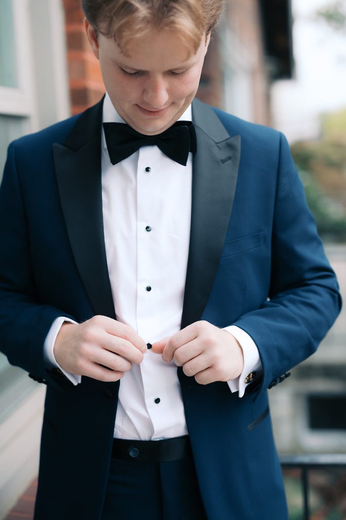 Groom adjusts his tuxedo shirt buttons while wearing navy blue suit with black bow tie