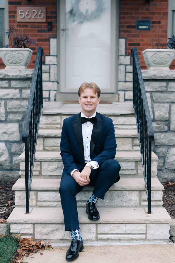 Groom Josh sits on stone steps in navy tuxedo with bow tie and patterned socks