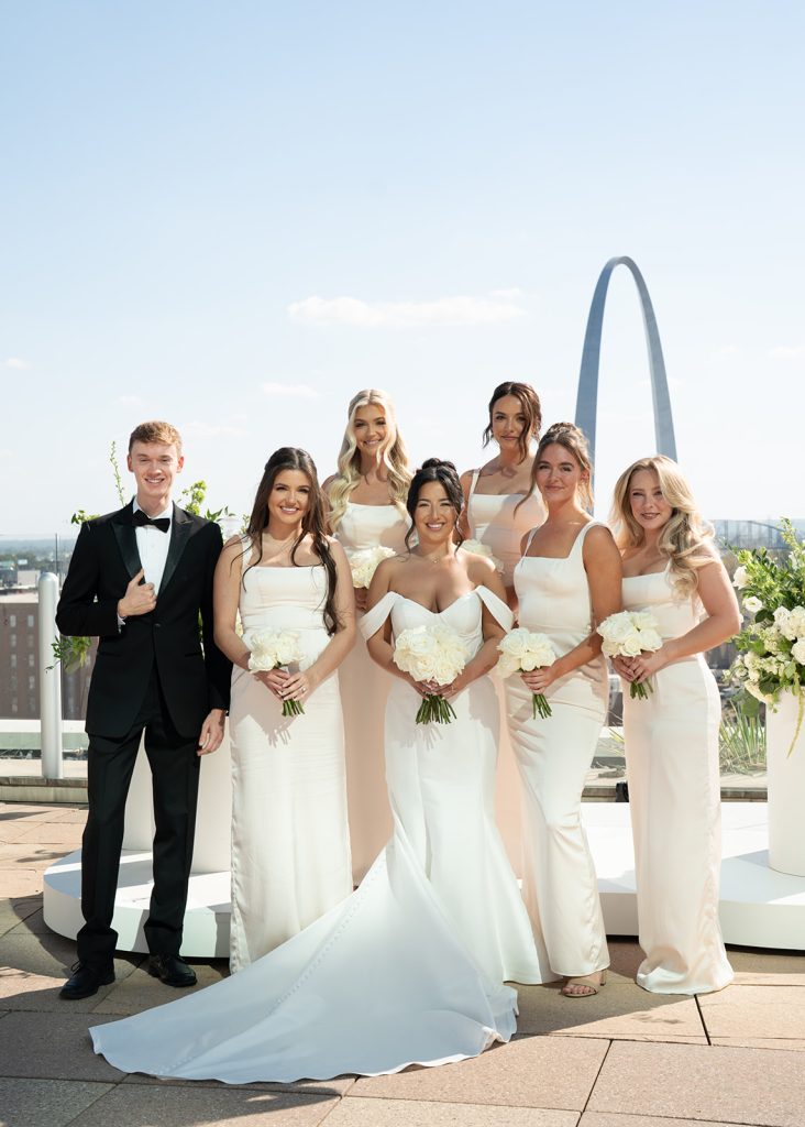 Katie and Anthony's bridal party with bridesmaids in ivory dresses and white bouquets pose on St. Louis rooftop with Gateway Arch skyline