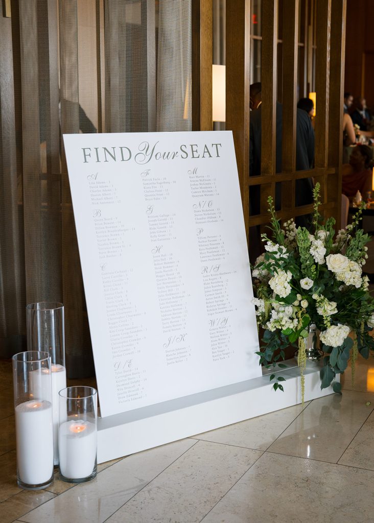 Wedding seating chart board flanked by pillar candles and white floral arrangement at reception entrance