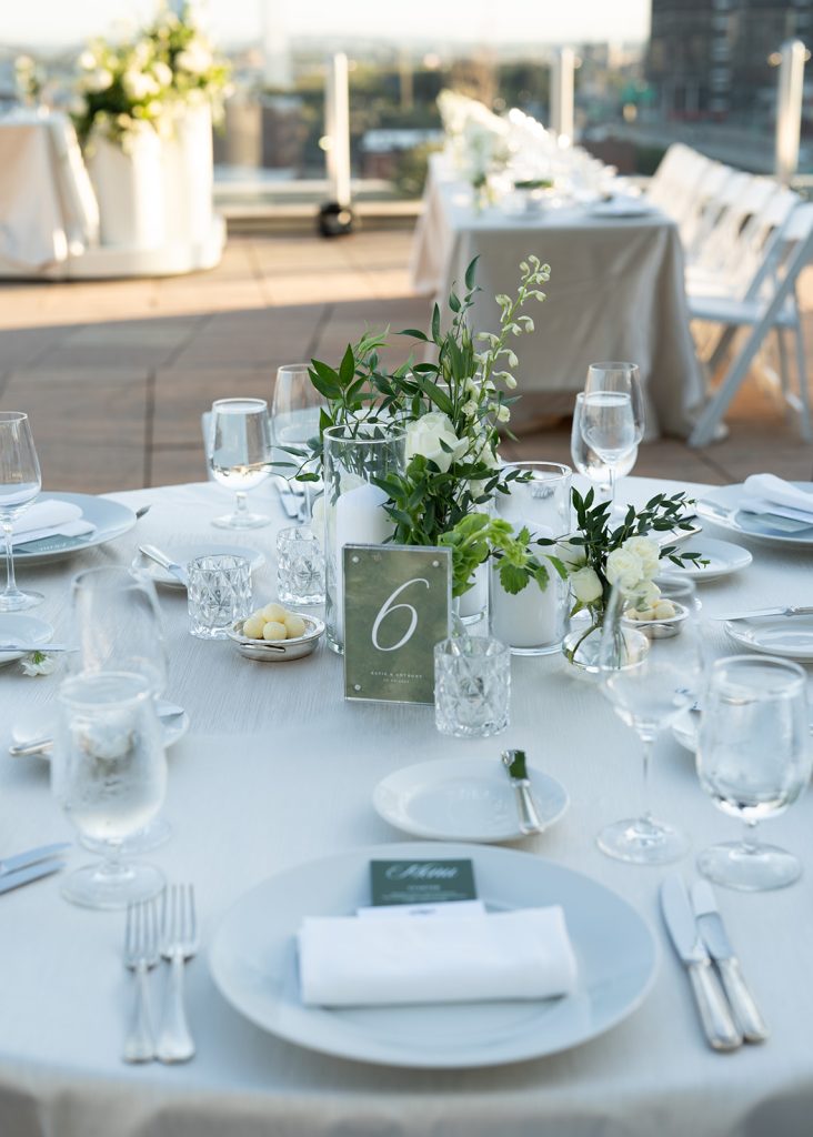 Rooftop wedding reception table setting with greenery centerpieces, table number six, and city skyline view