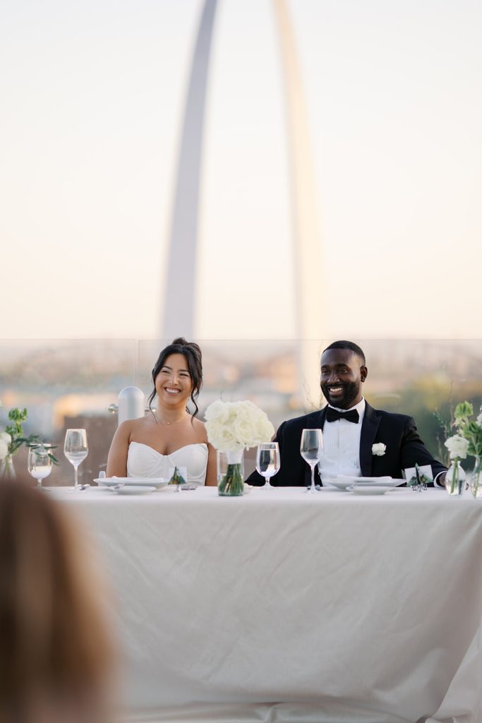 Katie and Anthony seated at their sweetheart table during golden hour with the Gateway Arch visible behind them