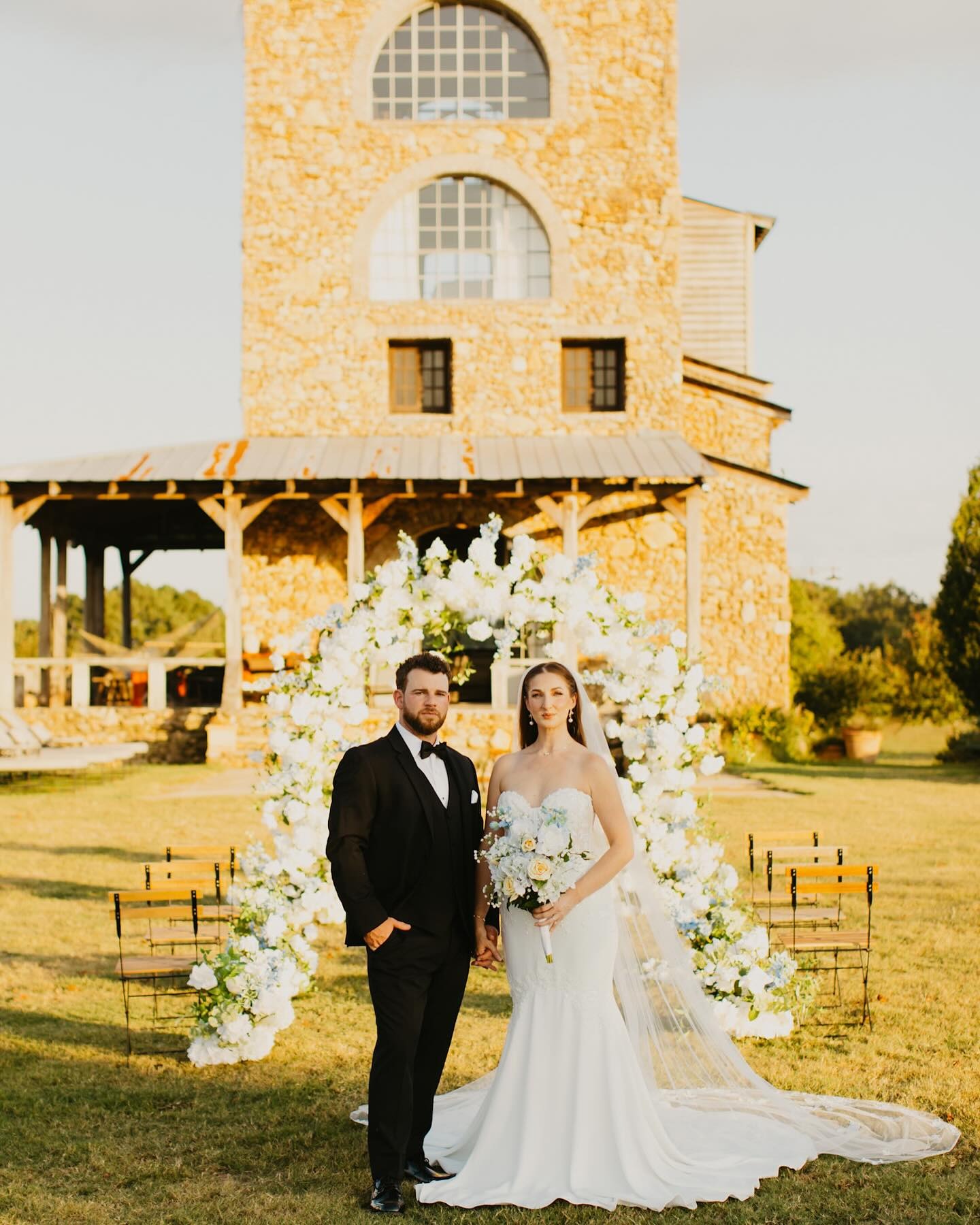 Bride in mermaid gown and groom in black tuxedo standing beneath white floral arch at rustic stone venue