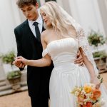 Groom in black tuxedo holds bride wearing ivory strapless dress and lace cathedral veil at outdoor venue