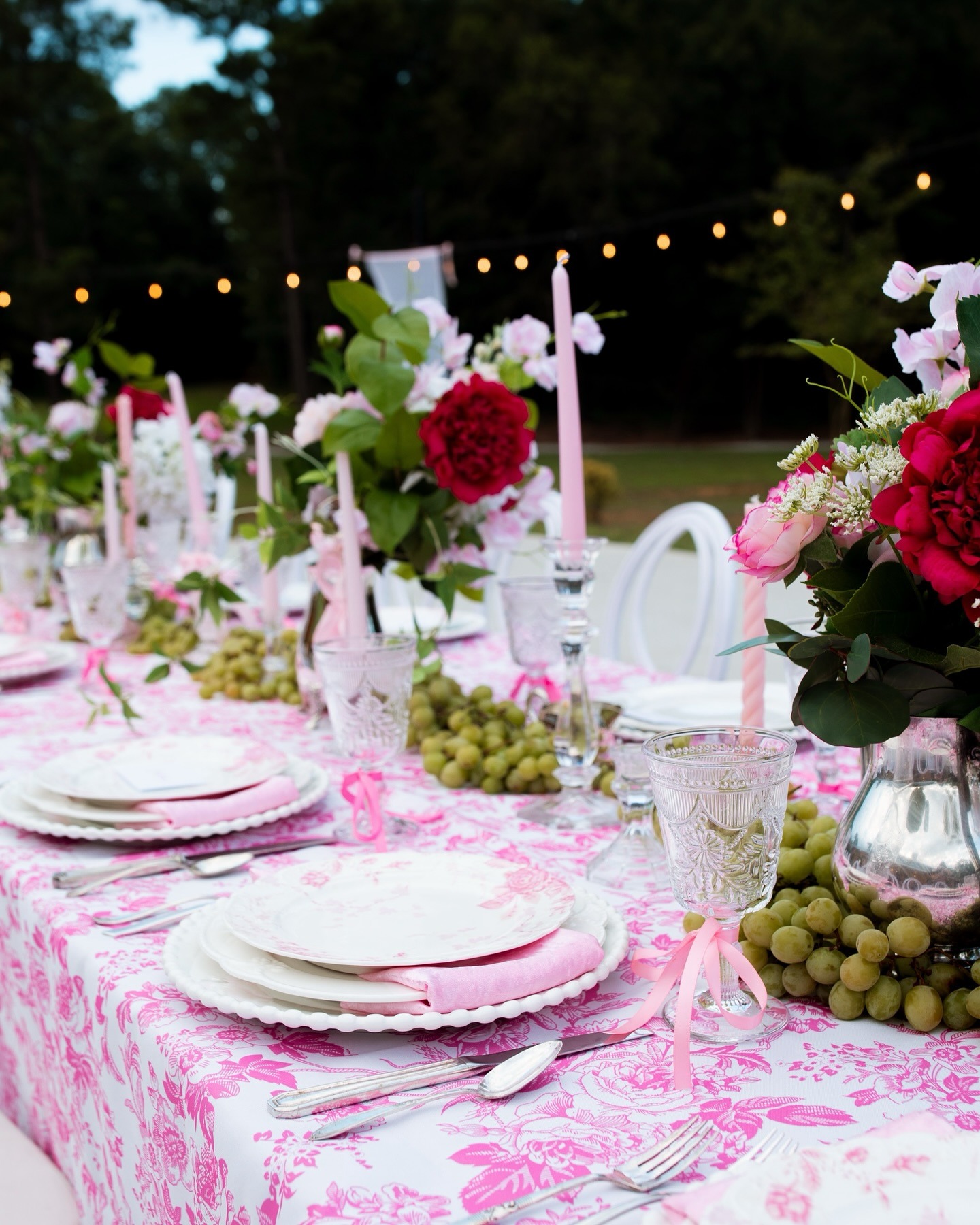 Evening wedding reception table with pink linens, illuminated by string lights and decorated with red and pink flowers