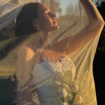 Bride in golden hour light holding veil above her head with white orchid bouquet, creating dramatic silhouette