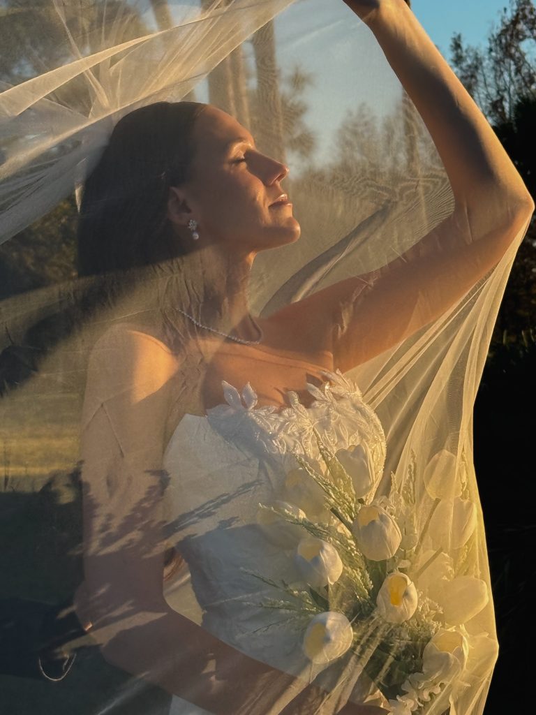 Bride in golden hour light holding veil above her head with white orchid bouquet, creating dramatic silhouette