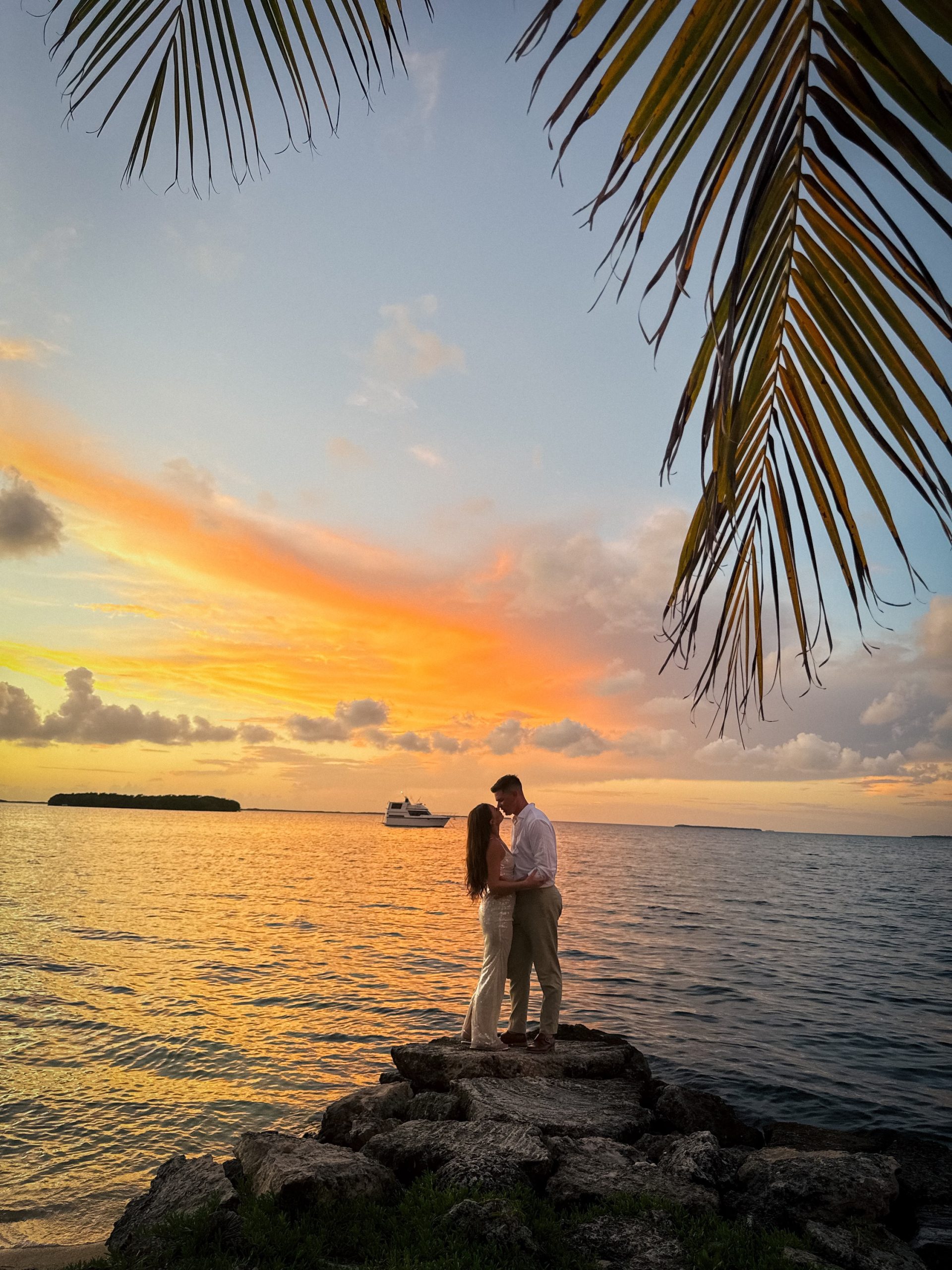 Couple silhouetted against vibrant sunset over water with palm fronds framing the scene