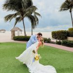Groom dips bride in romantic kiss on tropical beachfront lawn with palm trees and thatched huts