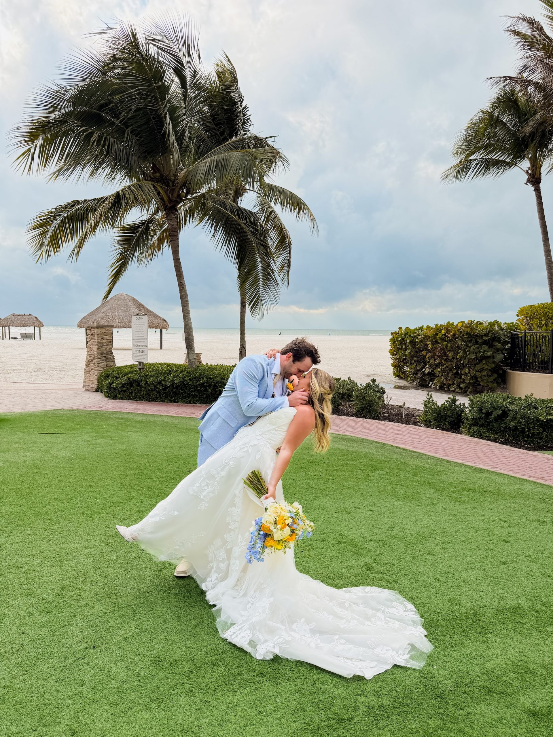 Groom dips bride in romantic kiss on tropical beachfront lawn with palm trees and thatched huts