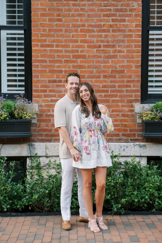 Engaged couple holding hands in front of red brick wall with black shutters and window boxes