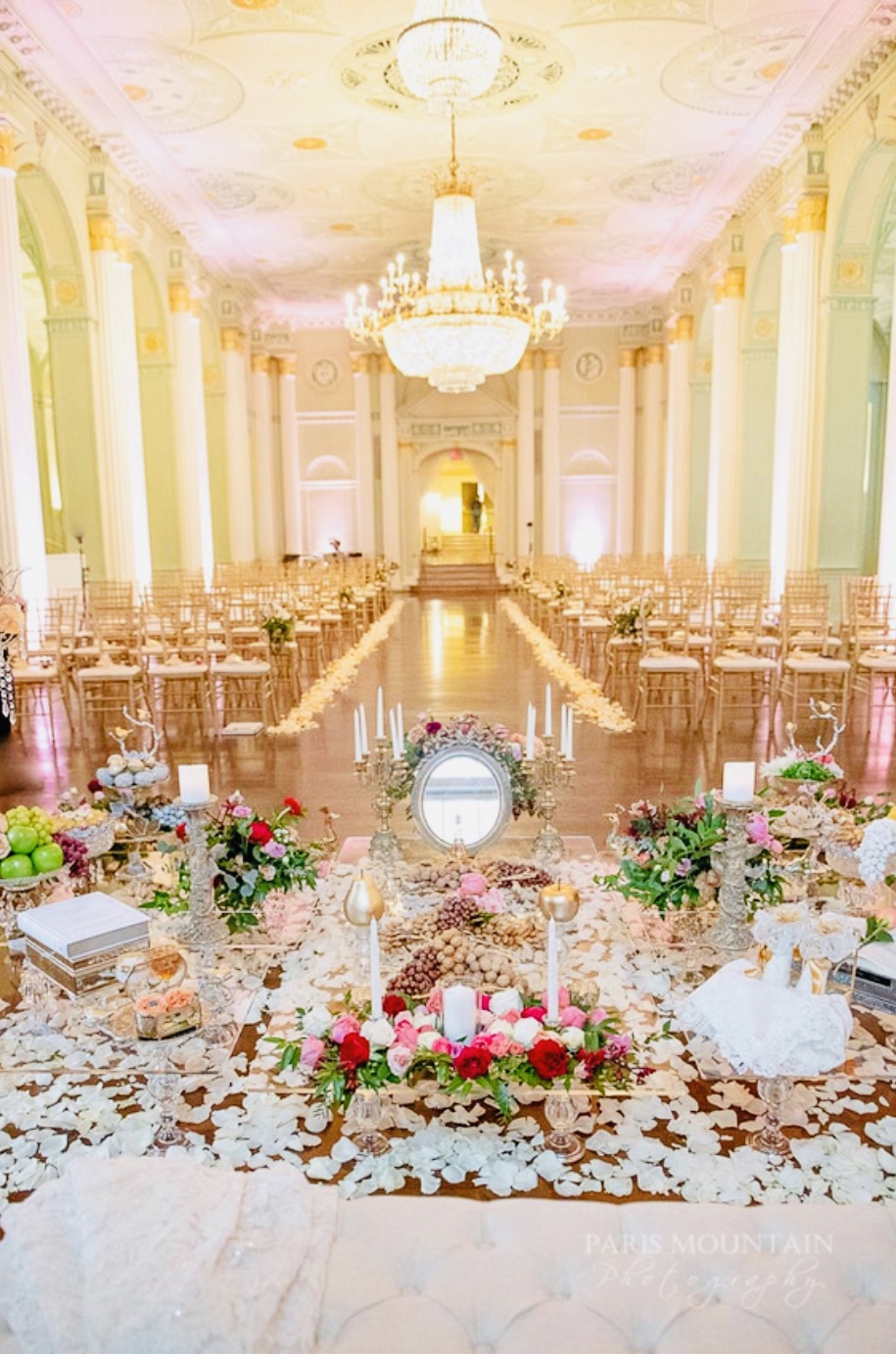 Elegant Persian Sofreh Aghd ceremony spread in grand ballroom with ornate chandelier, gold chiavari chairs, mirror, candles, nuts, and rose petals