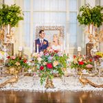 Couple standing behind ornate Persian wedding spread featuring colorful flowers, gold accents, and traditional ceremonial items