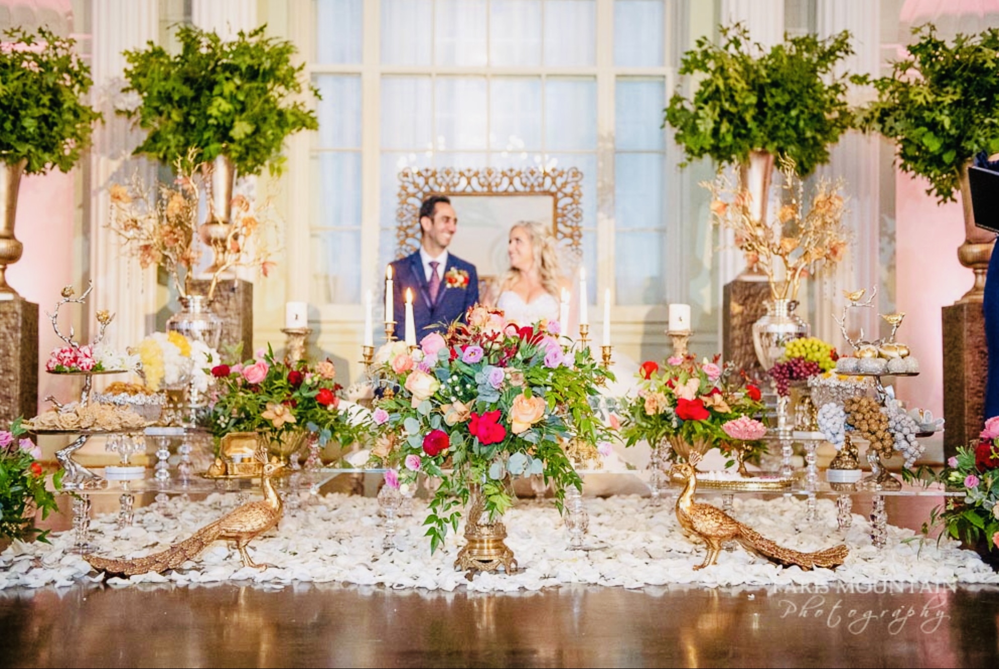 Couple standing behind ornate Persian wedding spread featuring colorful flowers, gold accents, and traditional ceremonial items