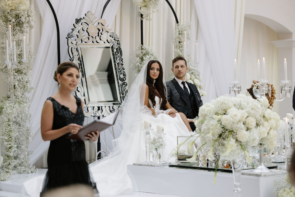 Bride and groom seated at white Sofreh Aghd ceremony table with officiant
