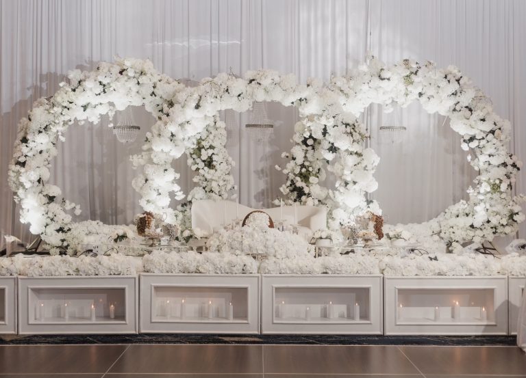 All-white Sofreh Aghd setup with dramatic floral arch, crystal chandeliers, and illuminated platform base