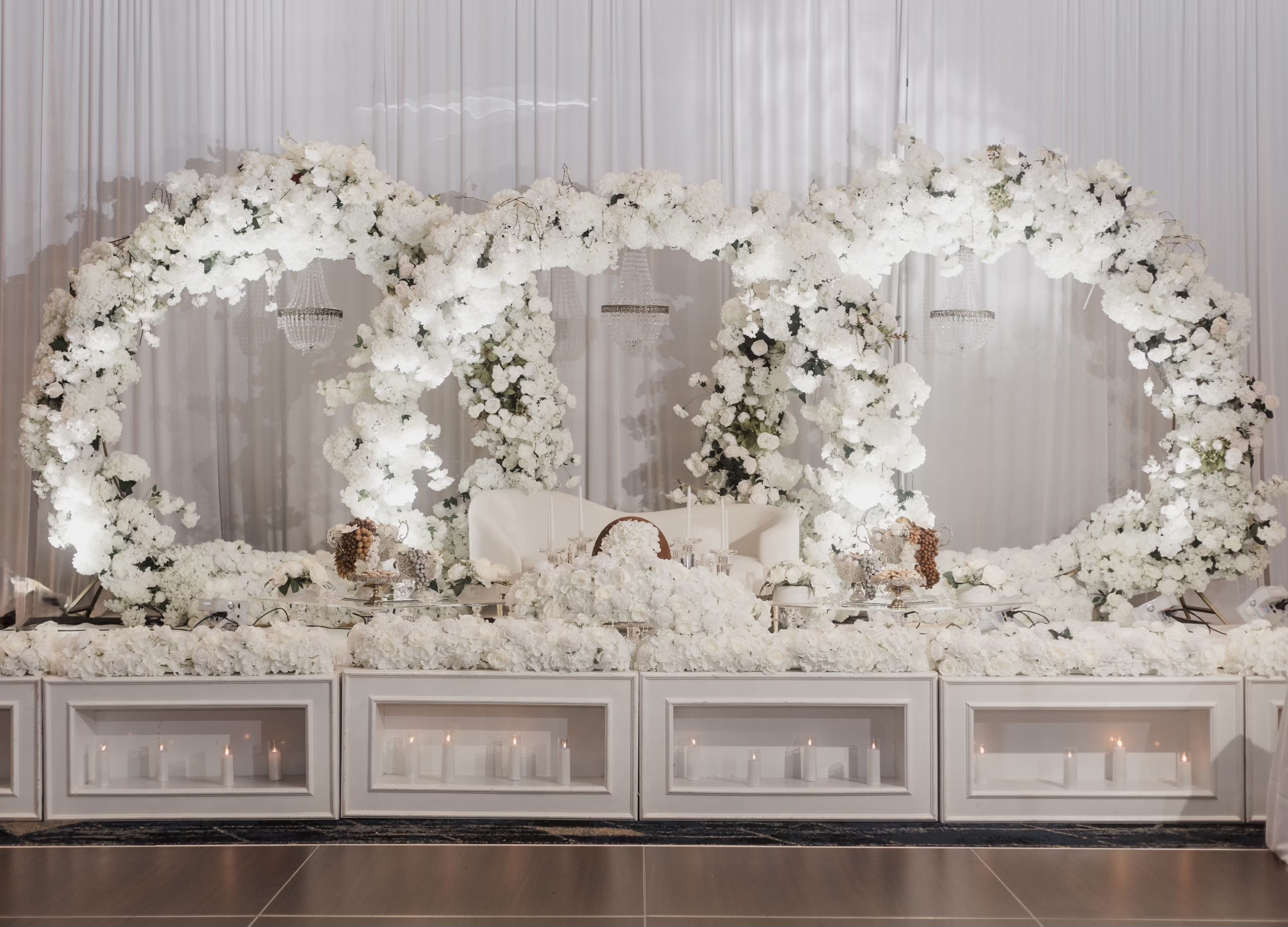 All-white Sofreh Aghd setup with dramatic floral arch, crystal chandeliers, and illuminated platform base
