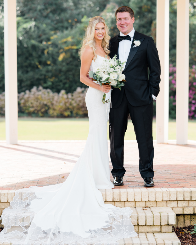 Bride in strapless white gown and groom in black tuxedo pose with white bouquet at outdoor wedding venue