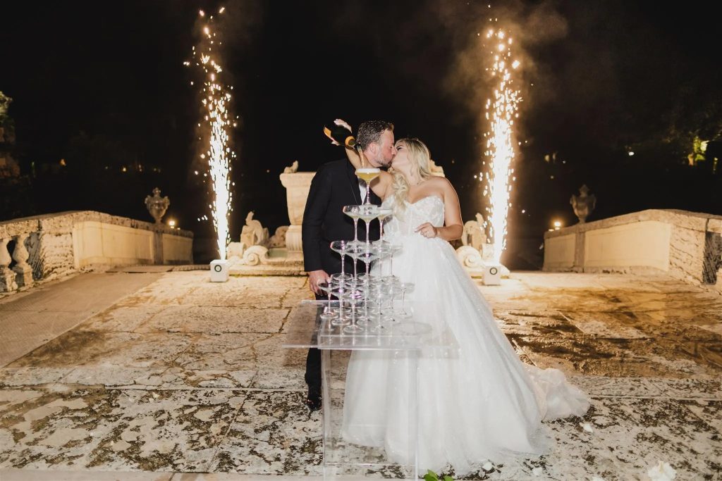 Bride and groom kissing behind champagne tower with dramatic sparkler fountains at outdoor nighttime reception