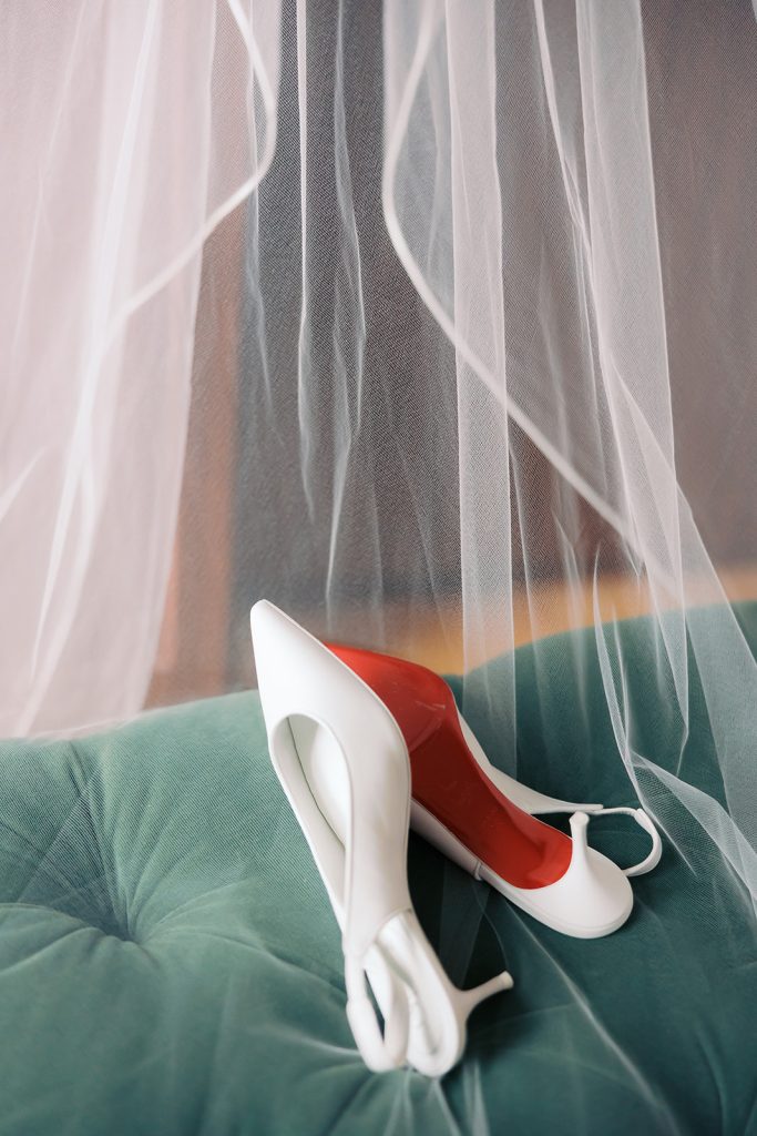 White bridal heels with red soles rest on teal velvet chair beneath flowing wedding veil