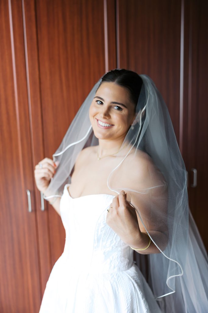 Caroline smiles in her strapless wedding gown and veil against warm wooden closet doors