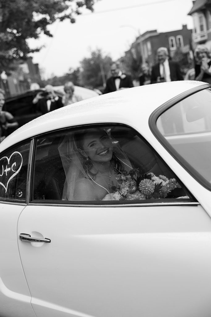 Bride Caroline beams from vintage white getaway car with wedding bouquet and "C+J" window decoration