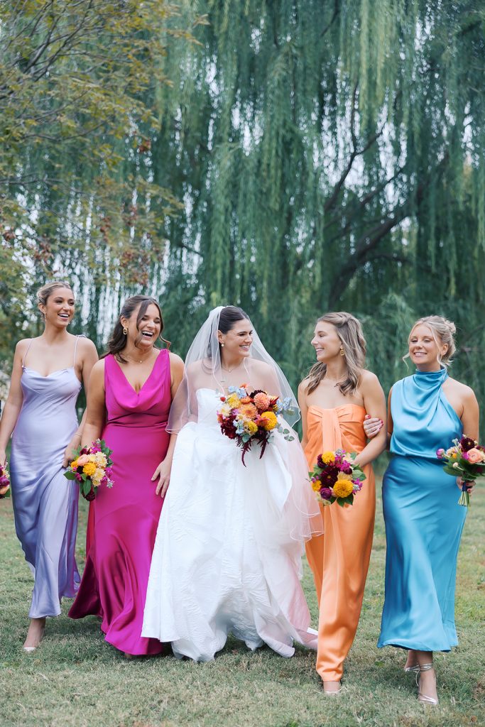 Bride Caroline with four bridesmaids in vibrant jewel-tone dresses—lavender, fuchsia, orange, and turquoise—beneath willow tree
