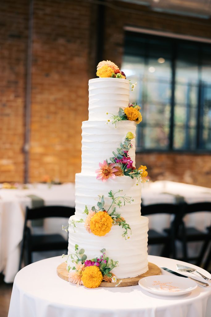 Five-tier white wedding cake decorated with vibrant yellow, pink, and burgundy fresh flowers cascading down the tiers