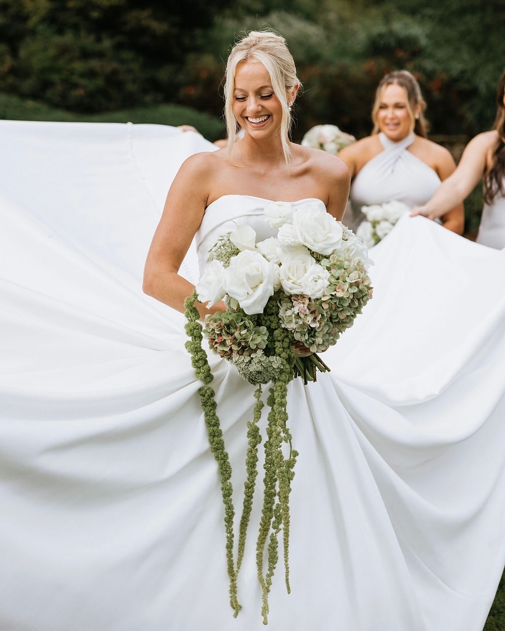 Bride holding oversized white and green bouquet with cascading amaranthus while bridesmaids look on
