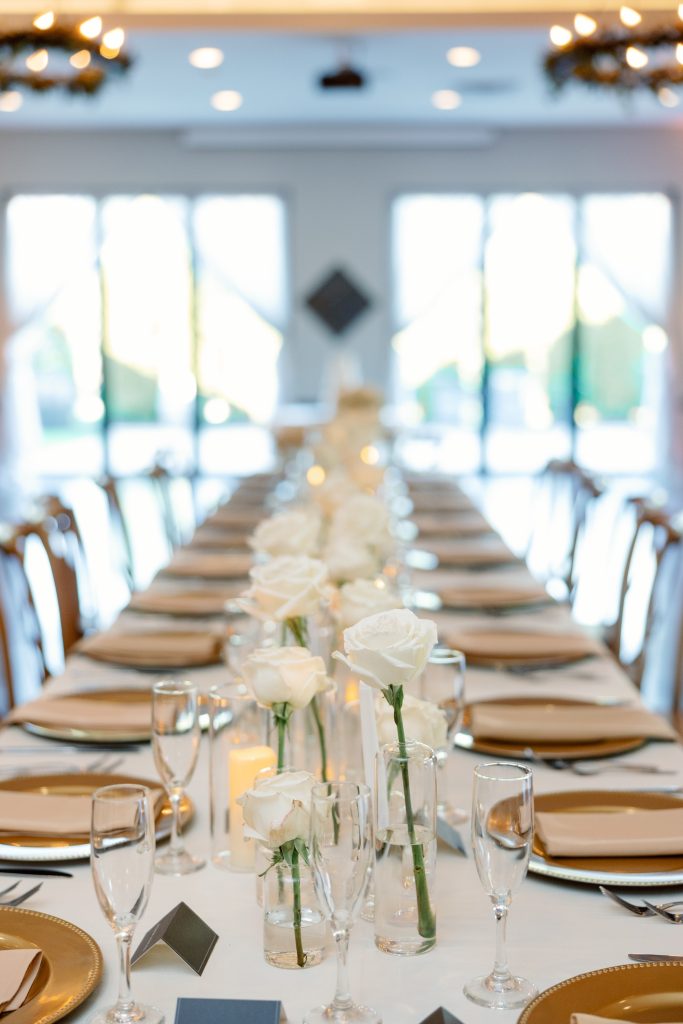 Long reception table with gold chargers, white roses in glass vases, and candles
