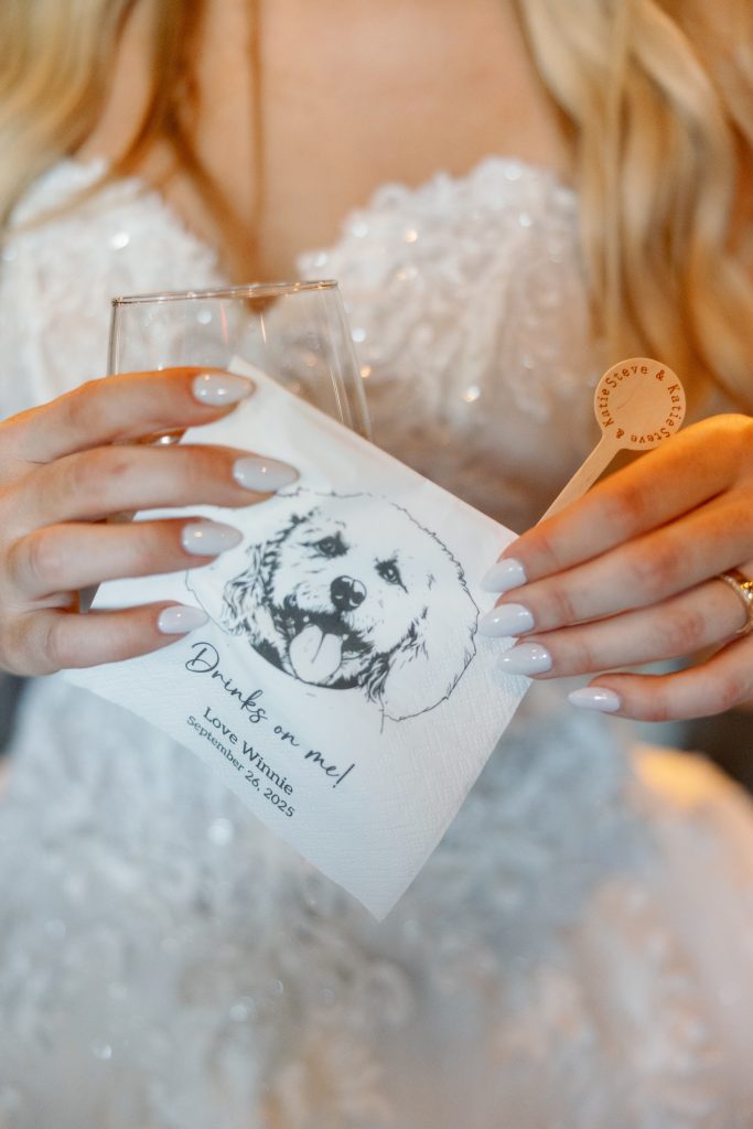 Bride holding custom cocktail napkin featuring illustration of golden retriever with "Drinks on me!" and Katie & Steve's wedding date