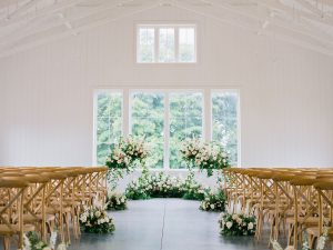 White barn wedding ceremony space with wood chairs arranged in rows and floral arrangements lining the aisle
