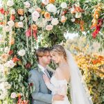 Couple kissing beneath vibrant floral arch featuring orange, pink, and white blooms