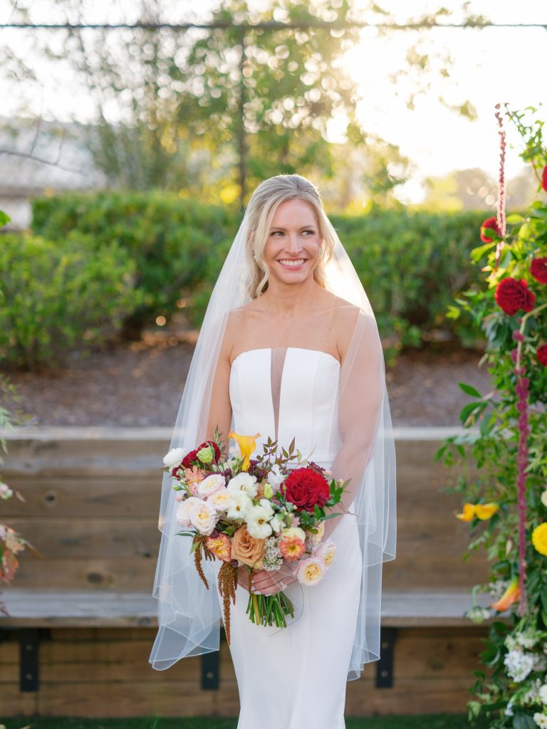 Smiling bride in strapless gown and cathedral veil holding lush bouquet with red dahlias, peach roses, and yellow blooms
