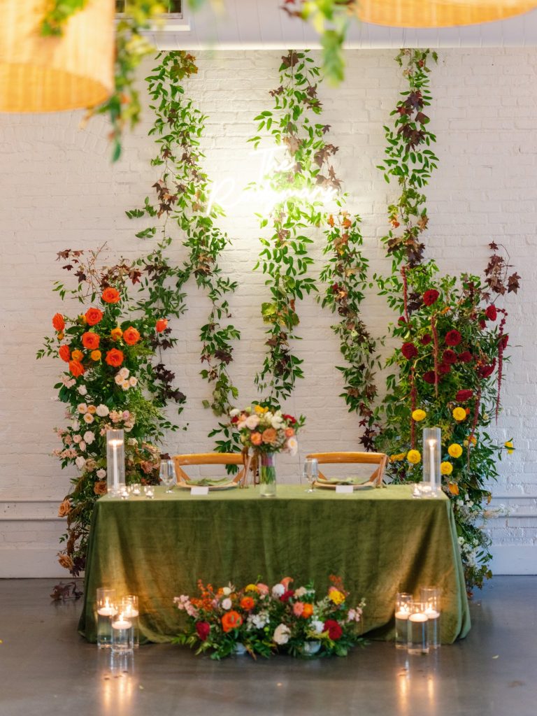 Sweetheart table with olive green velvet linens and white brick wall backdrop draped with cascading vines and multicolored roses