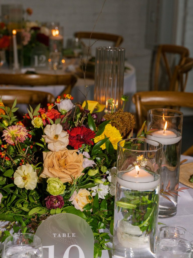 Colorful floral centerpiece with white, red, yellow, and burgundy blooms alongside floating candles and table number sign