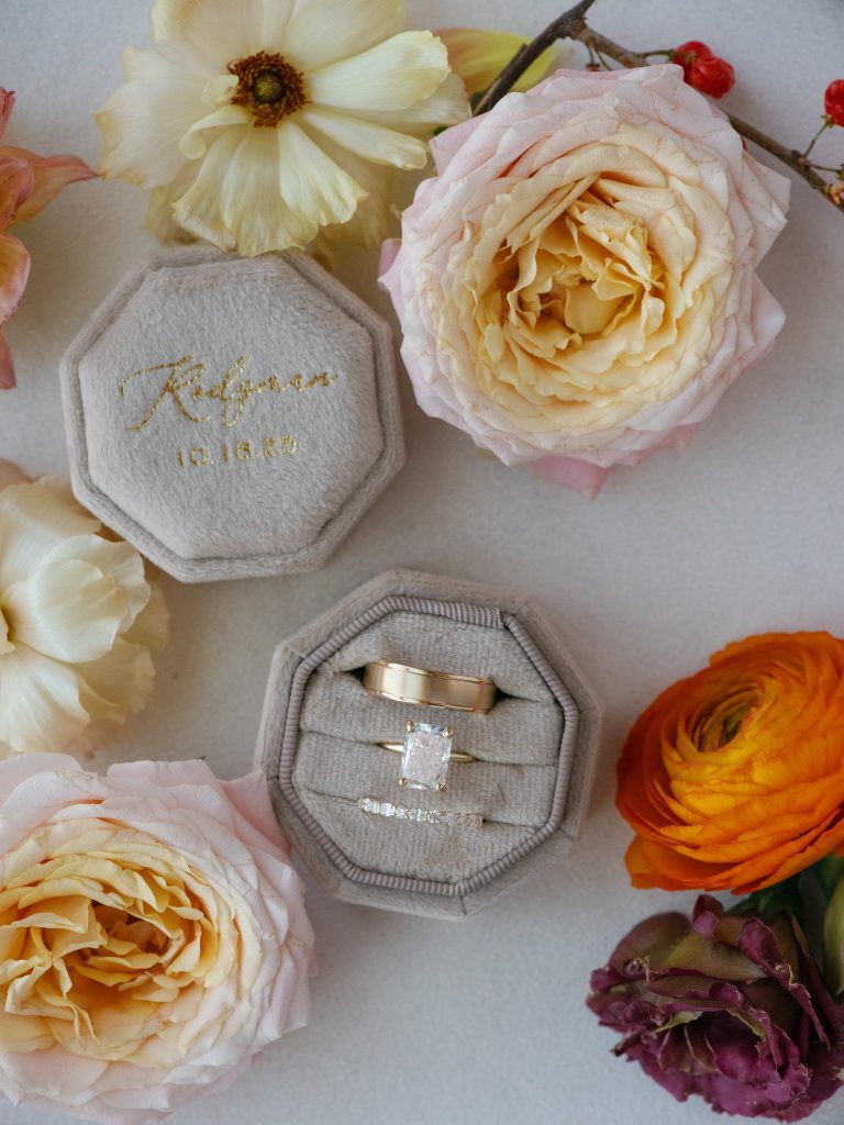 Wedding rings displayed in gray velvet box surrounded by cream, pink, yellow, and orange garden roses