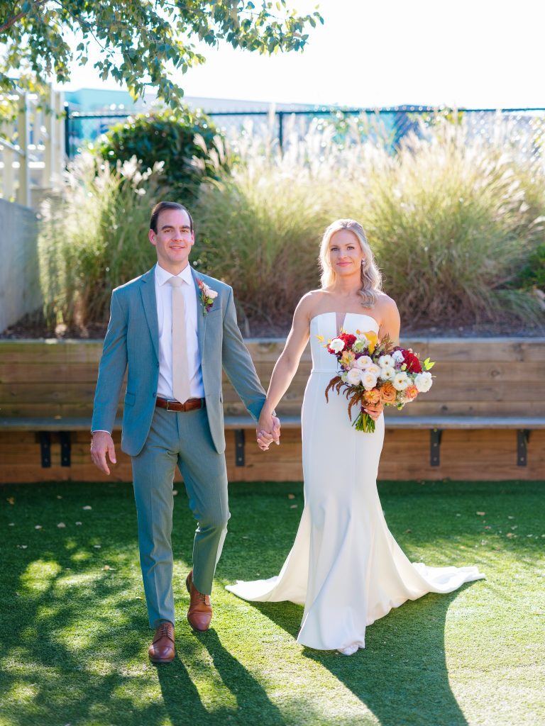 Newlyweds walking hand-in-hand across lawn, bride carrying vibrant multicolored bouquet with flowing trumpet gown train