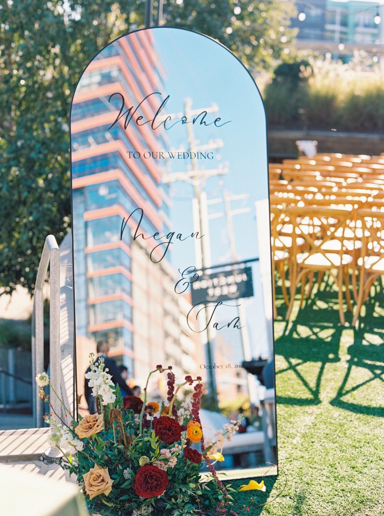 Acrylic welcome sign with couple's names reflecting urban downtown architecture, adorned with colorful florals at base