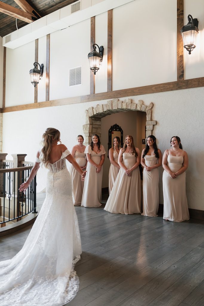 Bride in lace gown facing six bridesmaids in champagne-colored dresses in rustic venue with exposed beams