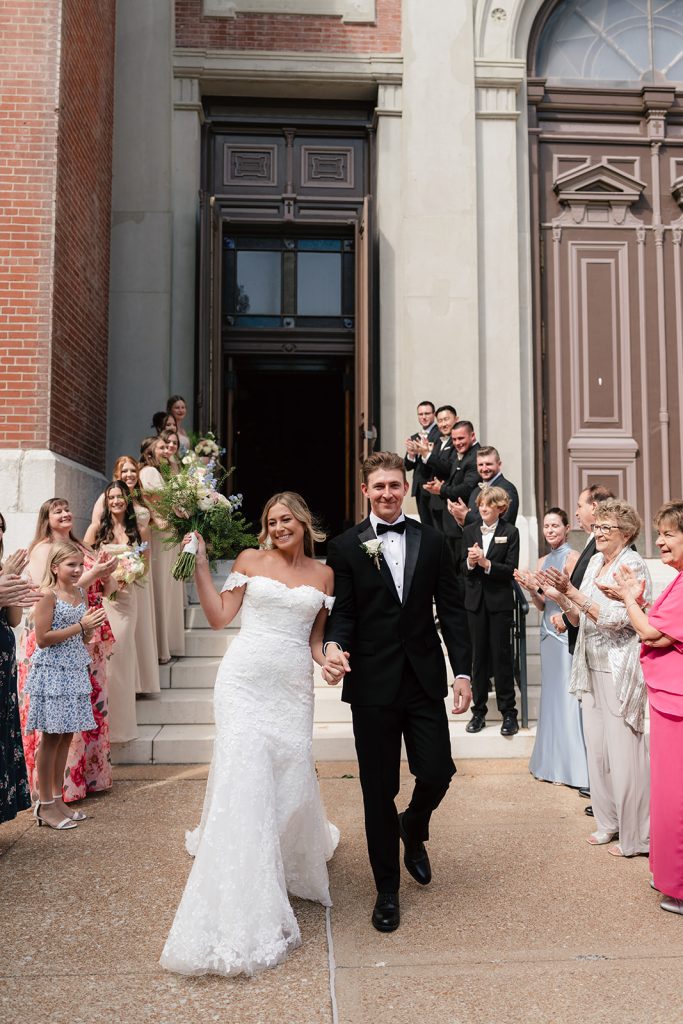 Rachel and Aaron exit their wedding ceremony as guests applaud and shower them with flower petals on church steps