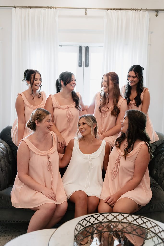 Bride in white slip dress surrounded by six bridesmaids in peach dresses, laughing together on black couch
