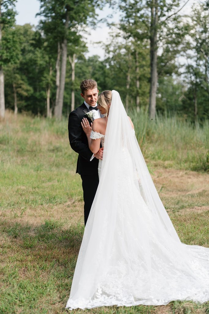 Rachel and Aaron embrace in open field with flowing cathedral veil and wooded backdrop