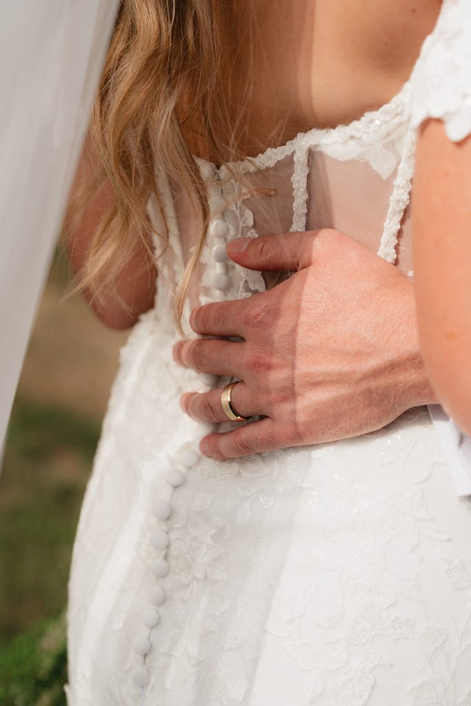 Close-up of groom's hand on bride's lace wedding dress back with illusion neckline detail