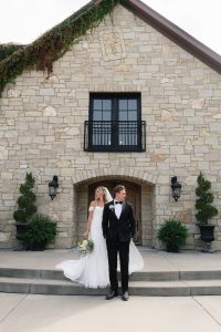 Caroline and Josh standing together outside a stone building with ivy-covered roof and black French doors