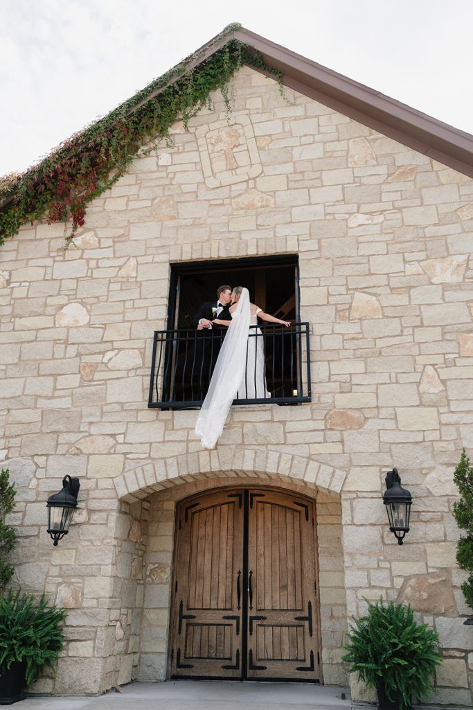 Newlyweds share a kiss on balcony of French-style stone building adorned with climbing ivy