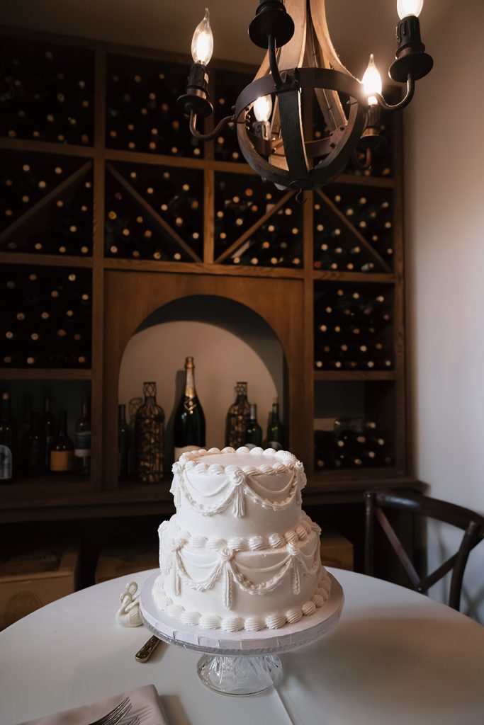 Two-tier white wedding cake with pearl garland details displayed on crystal stand in wine cellar with wooden chandelier