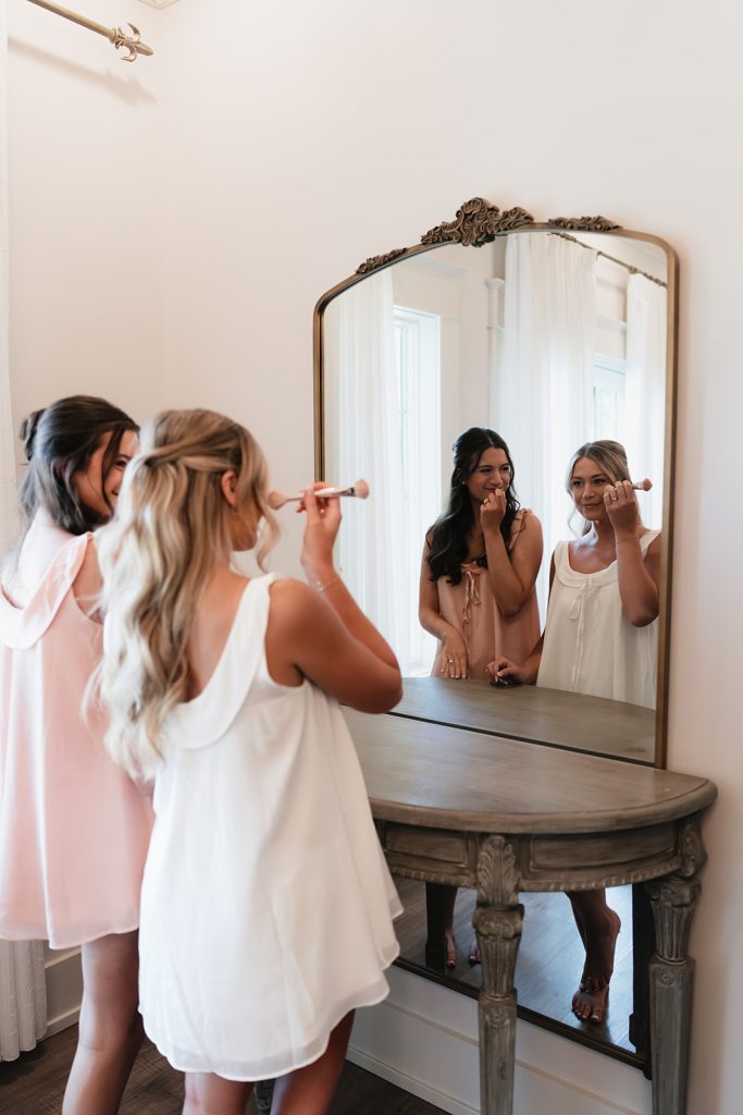 Bridesmaids apply makeup together in front of ornate gold-framed mirror during wedding preparations