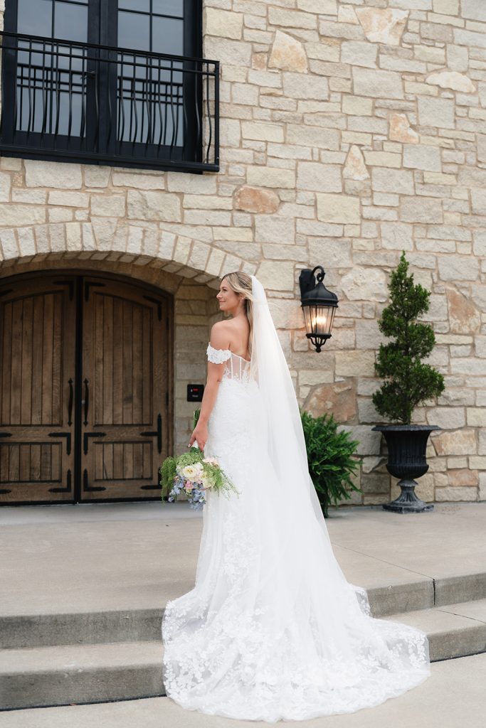 Bride Rachel poses in off-shoulder lace wedding gown with cathedral veil against French-inspired stone facade